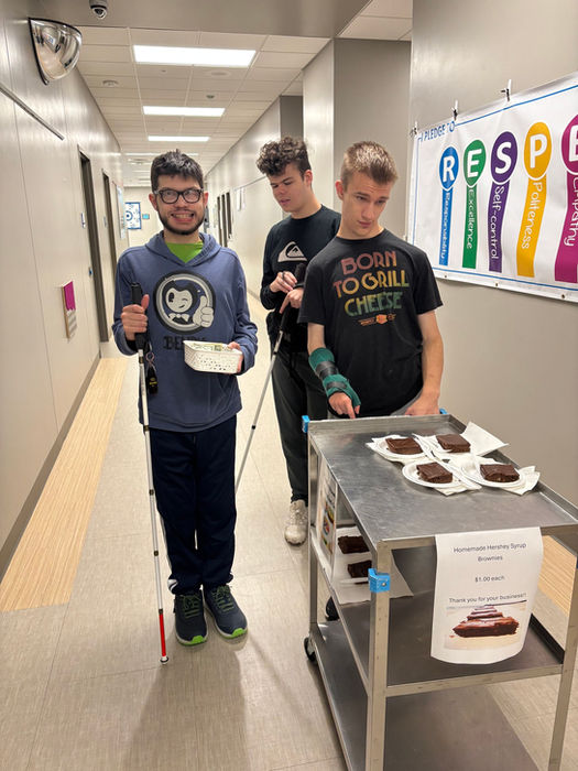 A group of three young men a standing behind a cart filled with individual brownies to sell around school as part of their small business project