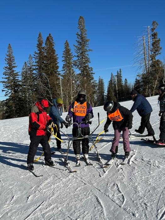 A young man on downhill skis holding onto a slider device for balance, standing with his guides after a fun run down the hill at Ski for Light in Black Hills, SD