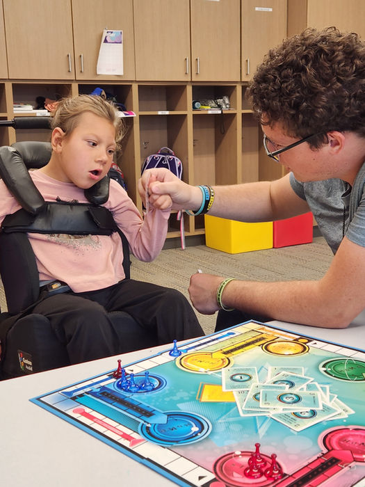 A young girl plays the game Sorry assisted by her Instructional Assistant