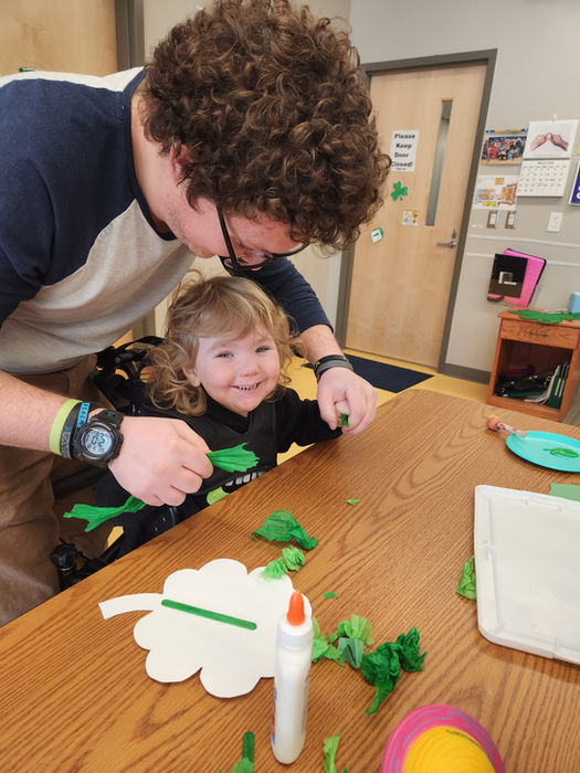 A young boy is smiling as he receives help from an instructional assistant with a green paper tissue craft
