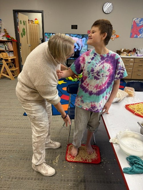 A woman leans beside a young woman who smiles while standing on dry macaroni