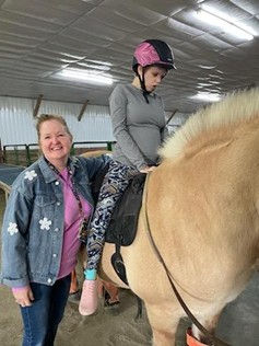 A woman smiles beside a horse while a young woman, wearing an equestrian helmet, sits in the saddle