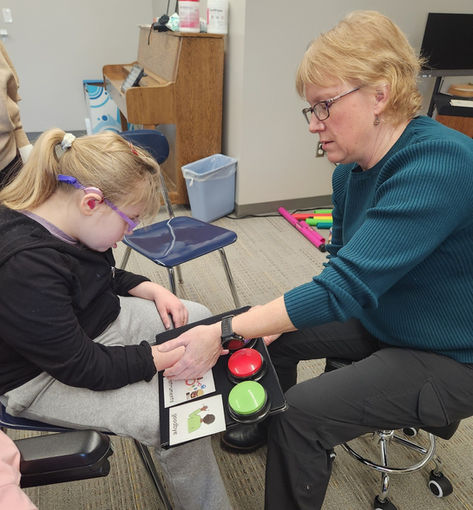 A Music Therapist uses picture cards labeled in Braille and Augmentative and Alternative Communication (AAC) buttons to show a young girl what activities will be performed next.