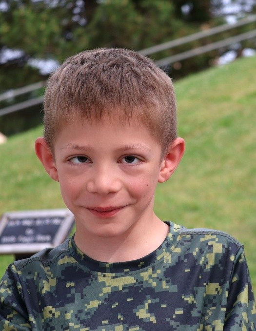 A young boy smiles at the camera with a large green hill in the background