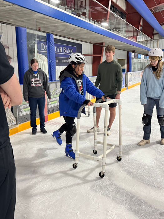 A young woman is ice skating with the aid of a wheeled walker while a crowd of students, a teacher, and Wings hockey players watch